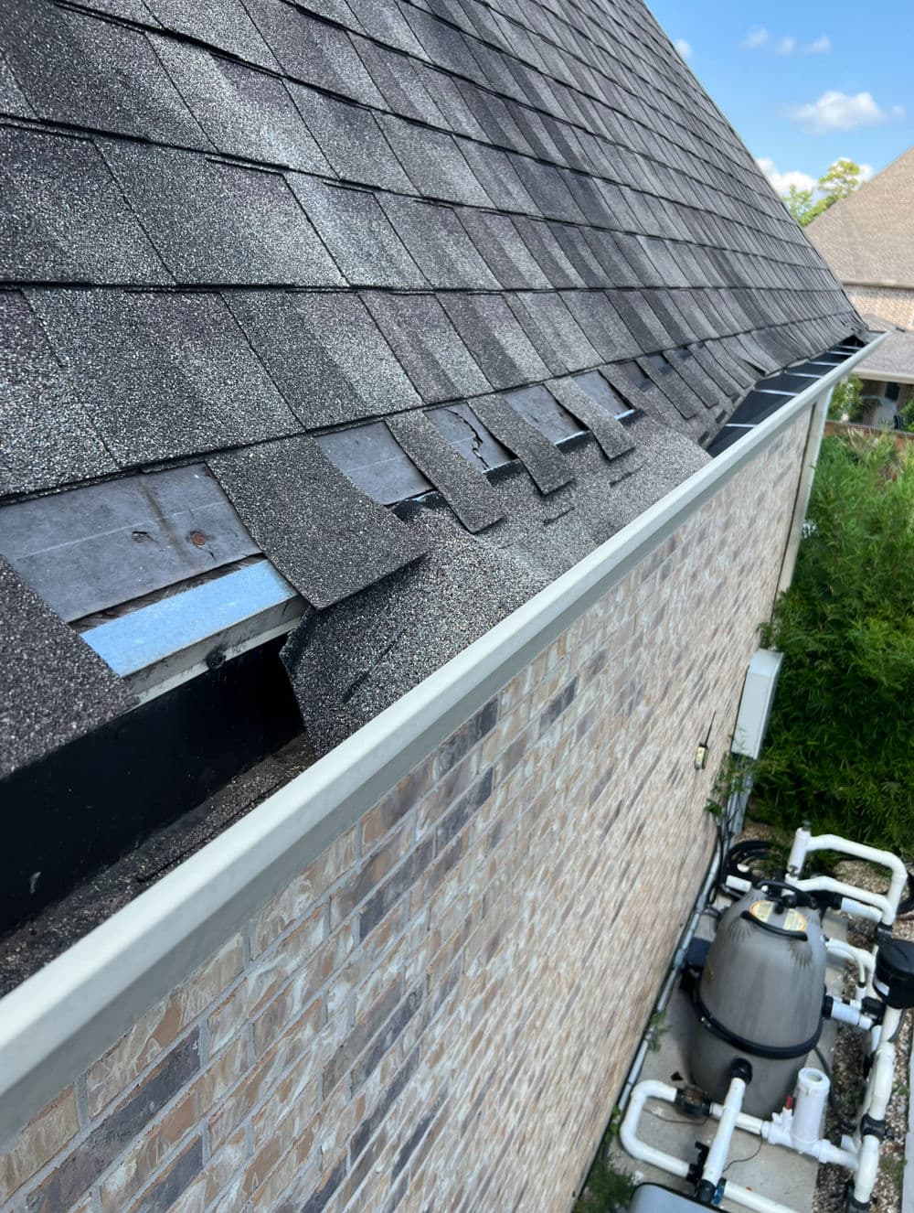 Damaged shingles on a house roof with gutter and pool equipment below.