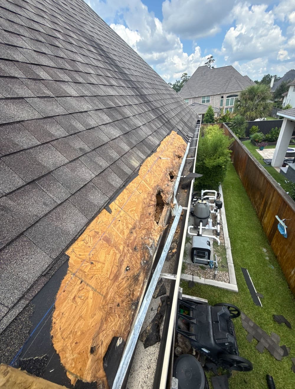 Damaged roof shingles exposing underlayment with cloudy sky background and backyard view.